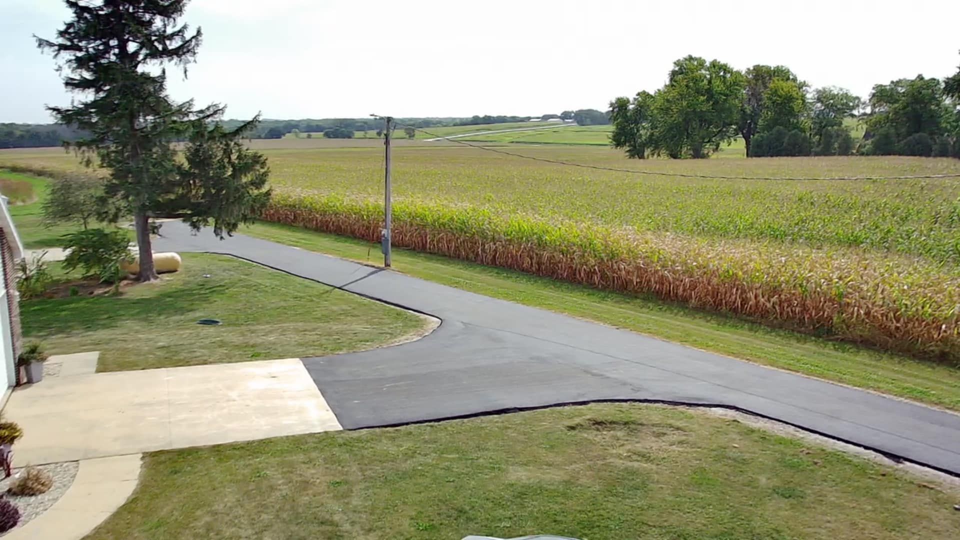 Paved driveway beside a grassy yard and cornfield under a cloudy sky