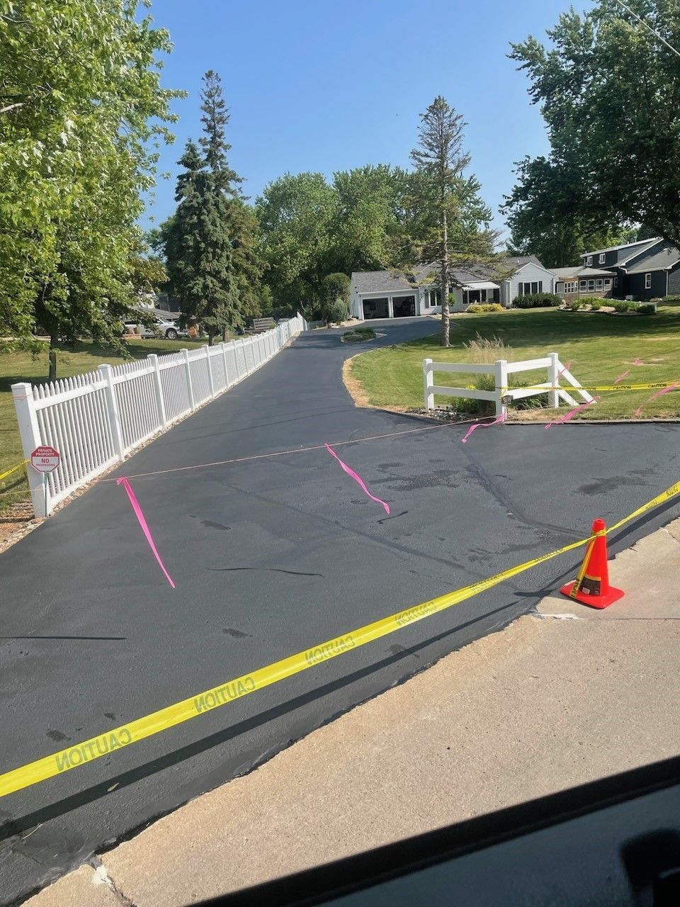 Driveway under construction with yellow caution tape, orange cone, pink markings, and a white fence beside a house.