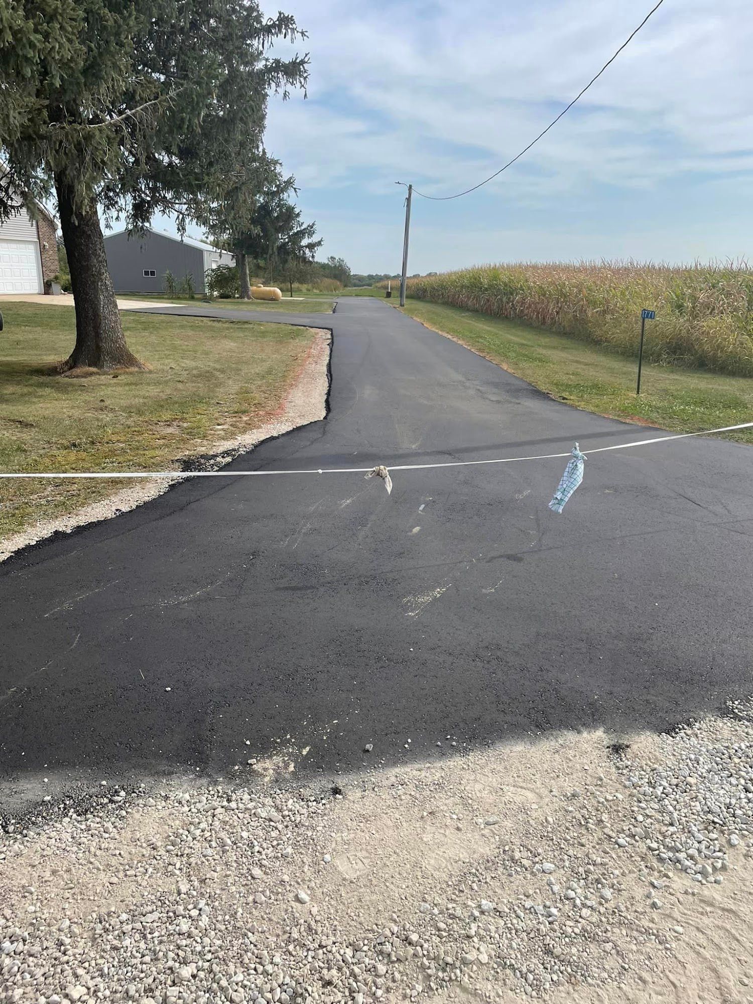 Rural paved road with a gravel shoulder, cornfield, and a tree-lined house on the left under clear sky.