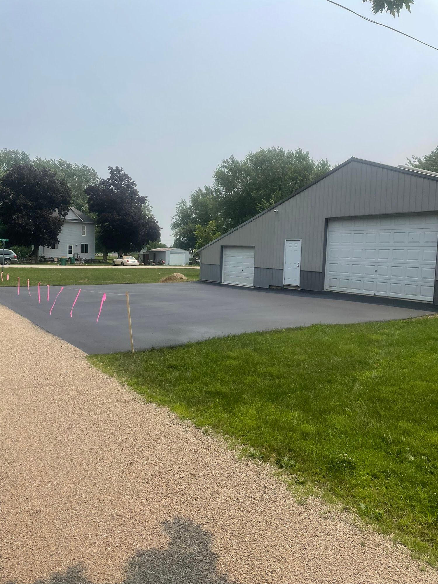 Paved lot beside a gray metal building with garage doors, gravel path, and trees in the background