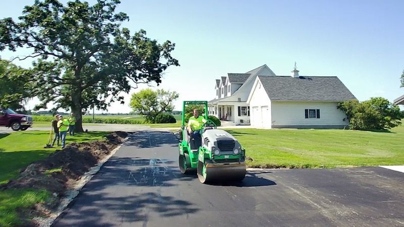 Green lawn mower on a paved driveway beside a white house and large tree on a sunny day