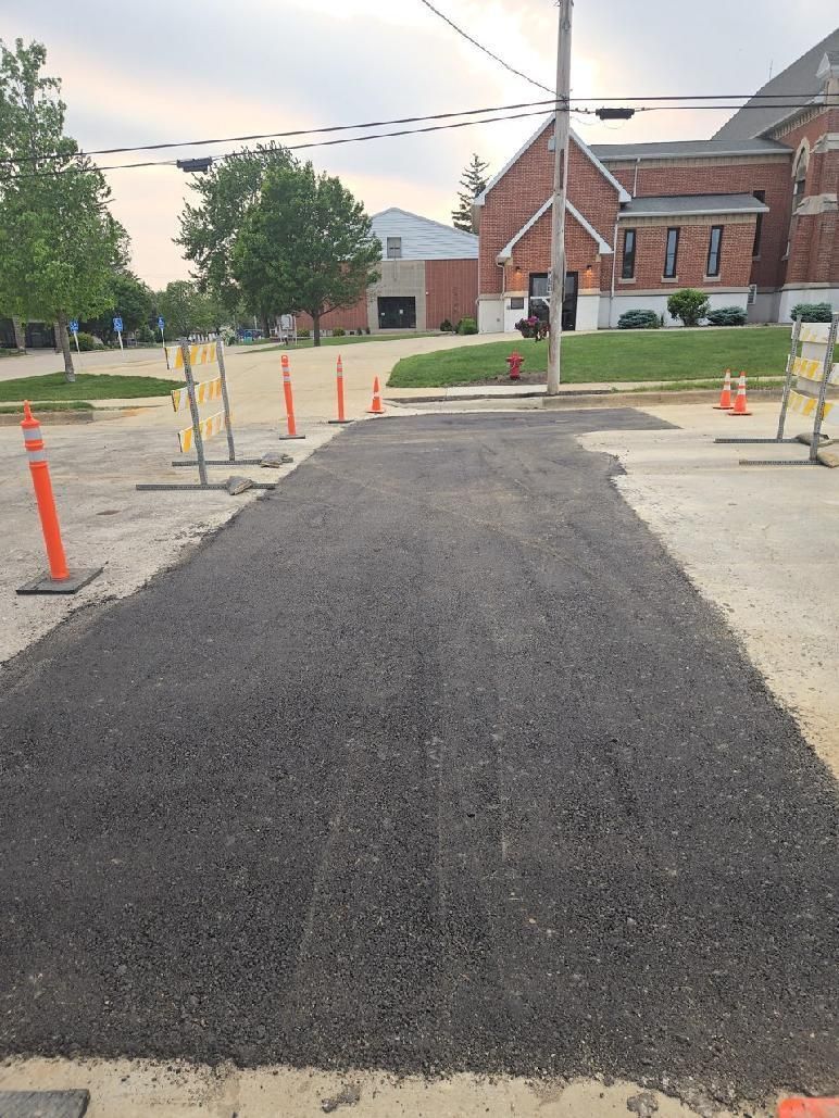 Fresh asphalt path under construction beside orange cones and a brick building