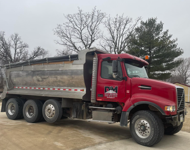 Red dump truck parked on a wet lot with bare trees in the background.