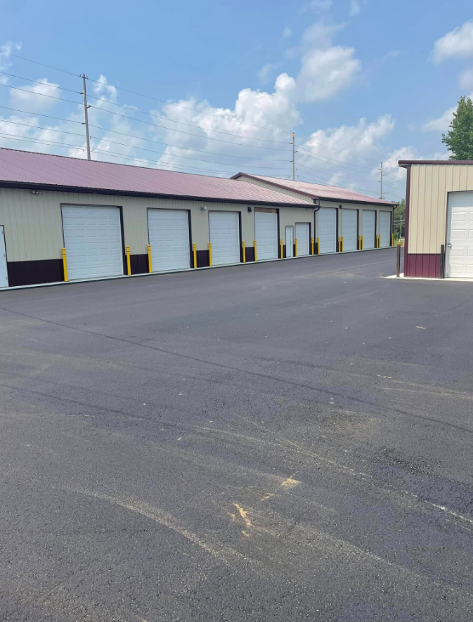 Industrial building with multiple white garage doors beside a large asphalt lot under a partly cloudy sky