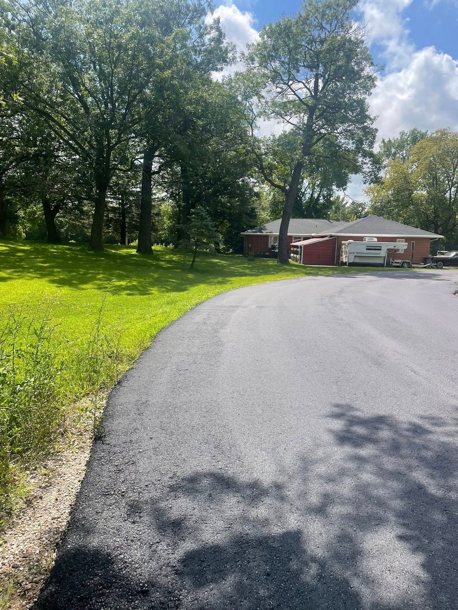 Curving paved driveway beside green lawn and trees, leading to a red house under a sunny blue sky.