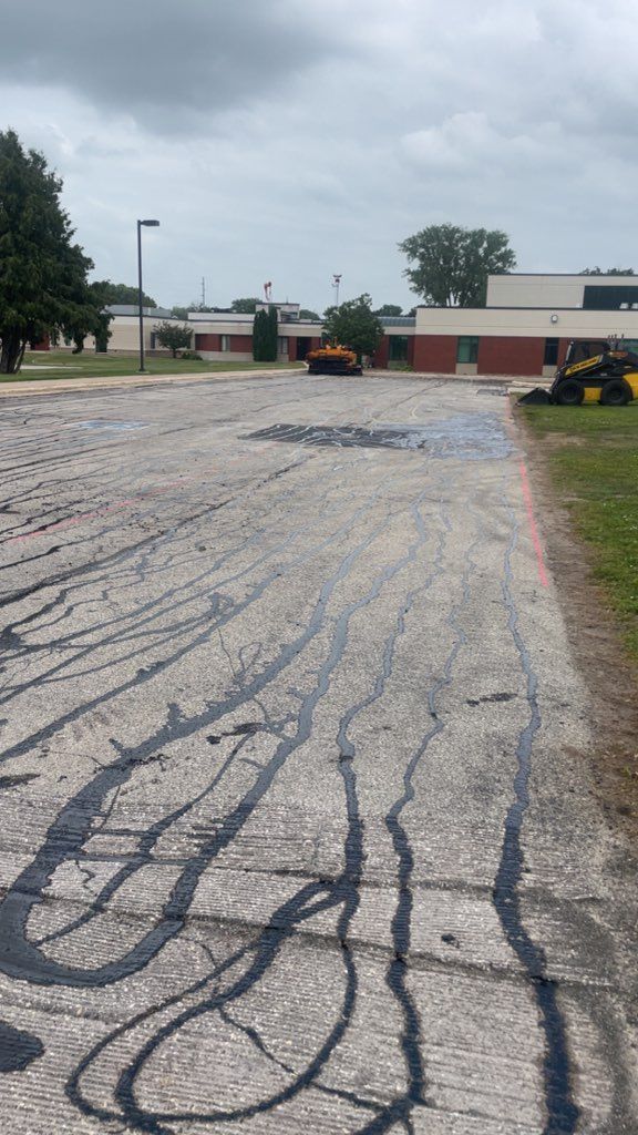 Cracked gravel parking lot beside low buildings under a cloudy sky, with a small construction vehicle in the distance