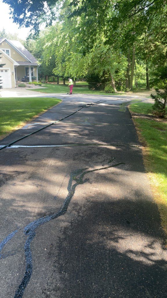 Cracked residential driveway shaded by trees, with a house on the left and a person in the distance.