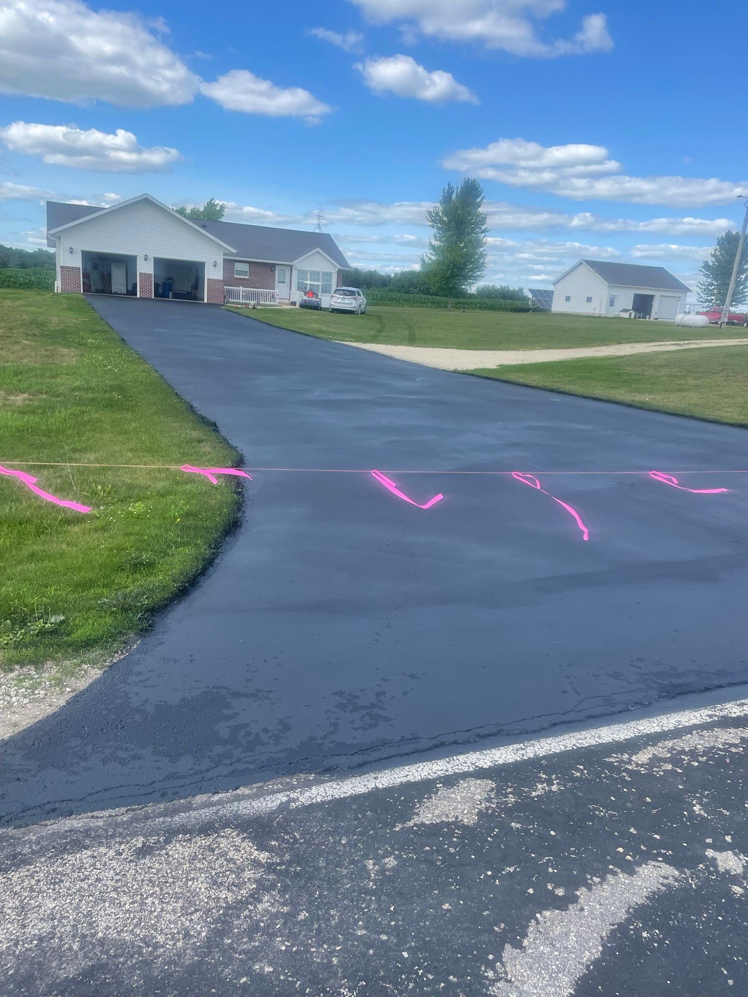 Fresh black asphalt driveway curving up to white houses under a blue sky, with pink markings on the pavement