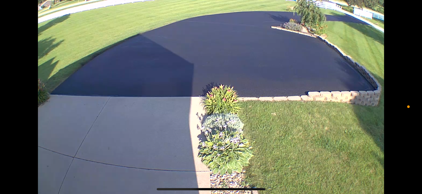 Driveway and front lawn beside a large dark asphalt area, with a small flower bed in the center.
