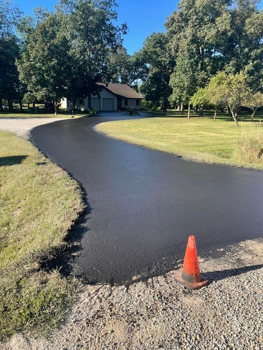 Freshly paved curved driveway beside a grassy yard, with an orange cone in the foreground and a house in the distance