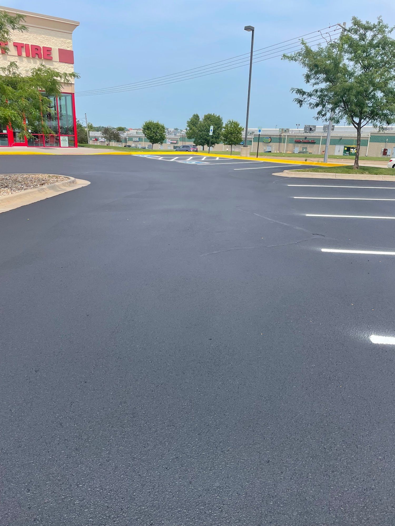 Empty parking lot with fresh black asphalt, yellow curb, trees, and a building at left under a cloudy sky
