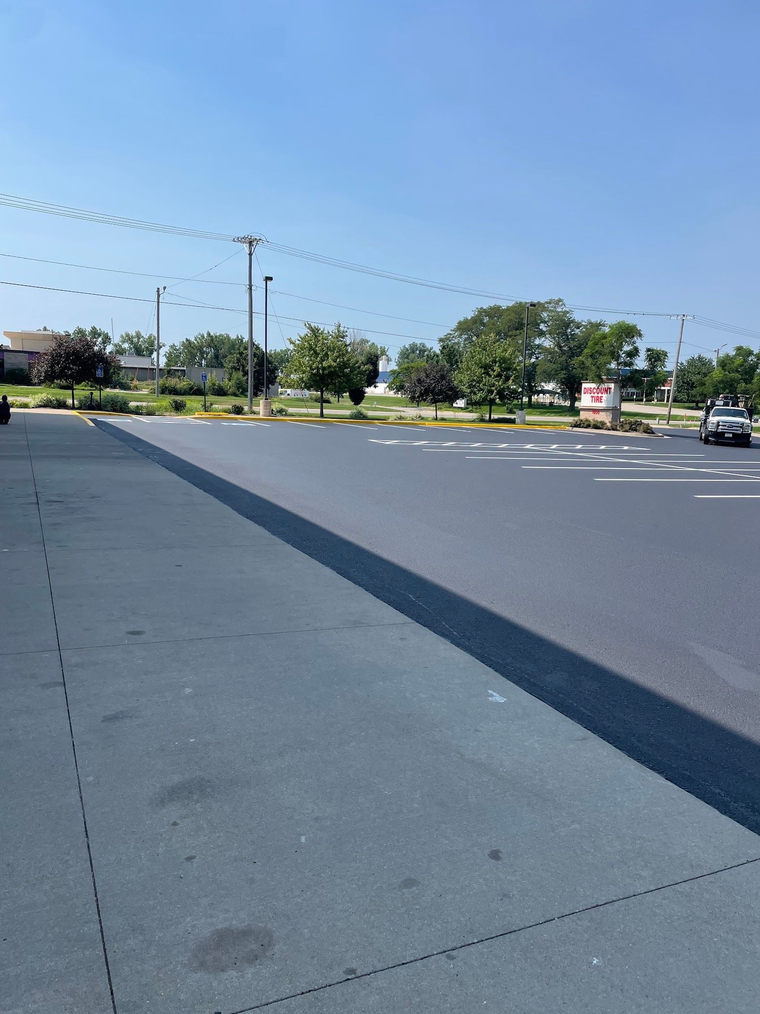Empty parking lot beside a road under a clear blue sky, with trees and a few cars in the distance.
