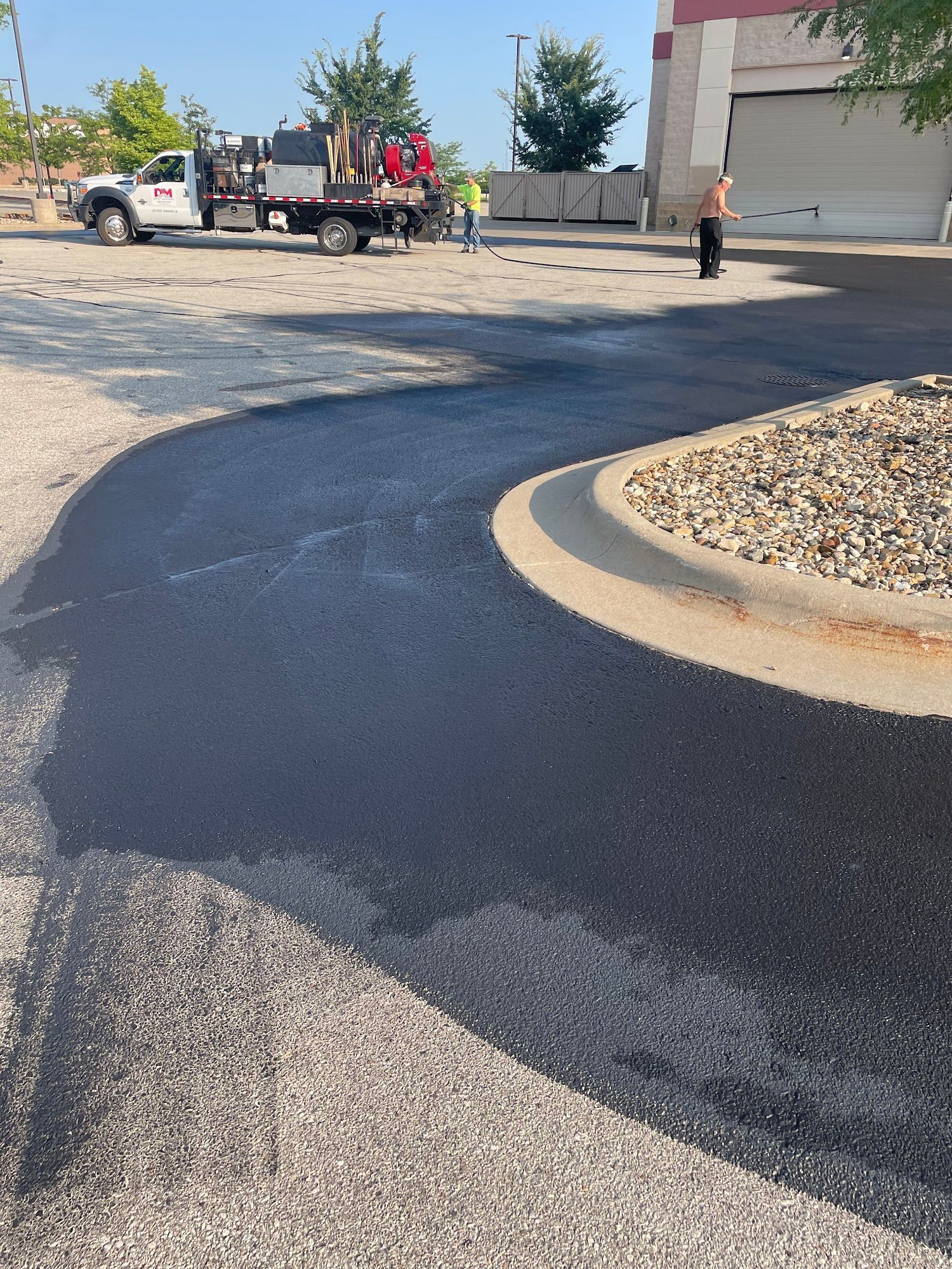 Freshly paved black asphalt driveway curving beside a gravel island near a building and work truck