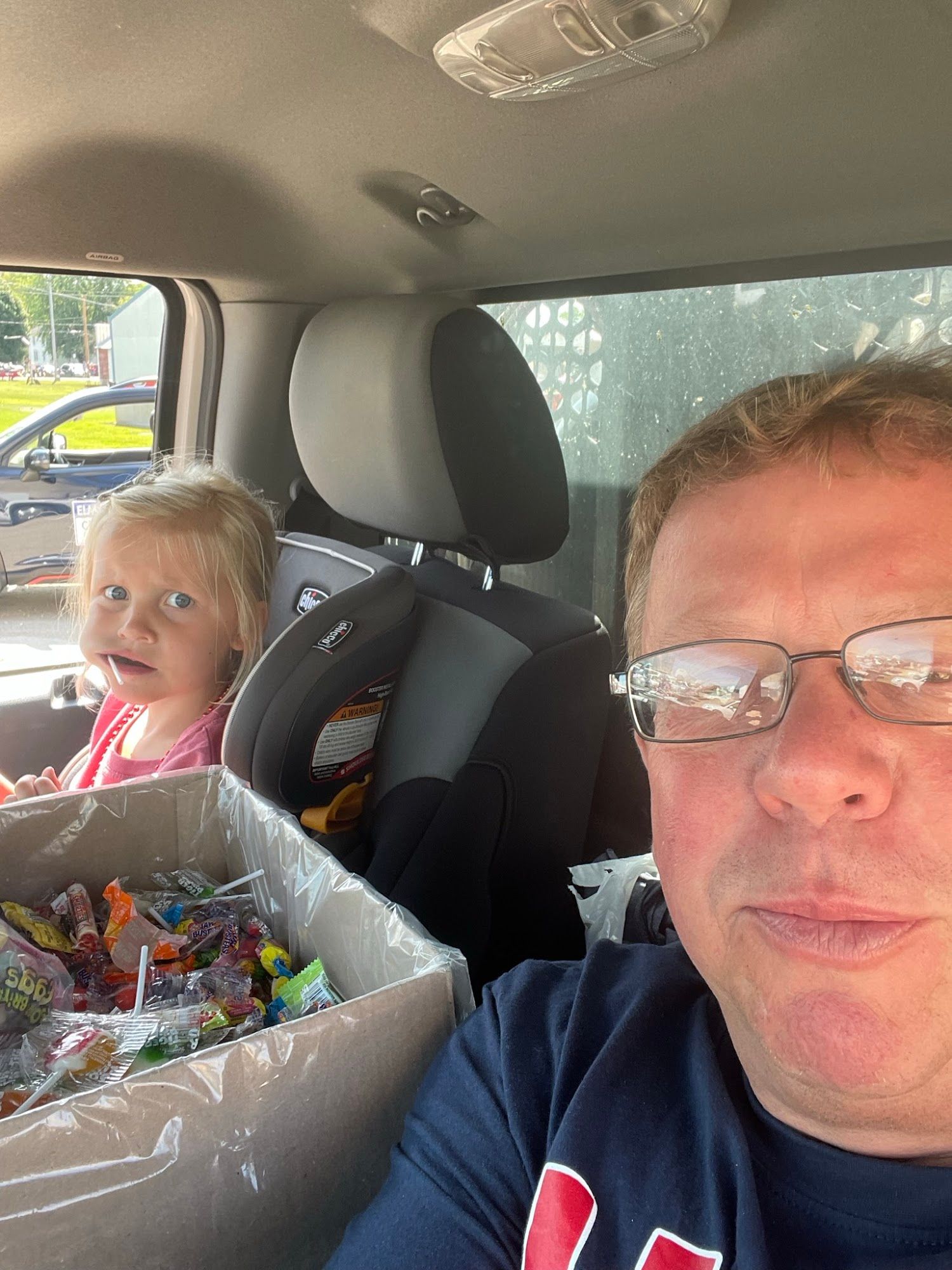 Two people in a car, smiling selfie with a child in the back seat beside a box of wrapped items