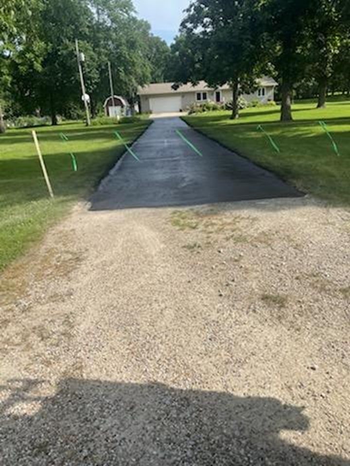 Gravel driveway leading to a house across a green lawn, with a wet black asphalt section in the middle.