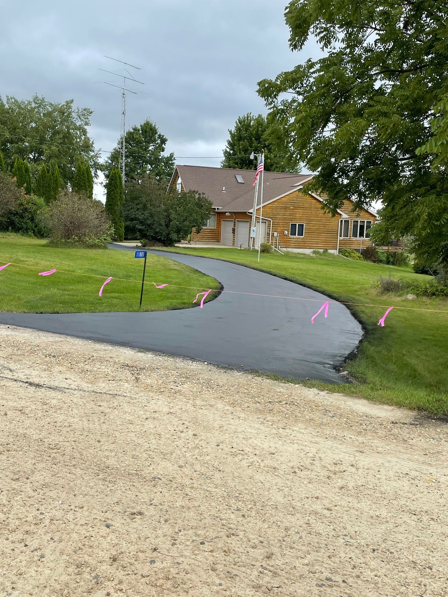 Curved new asphalt driveway beside a house and lawn, with pink survey markers under cloudy sky