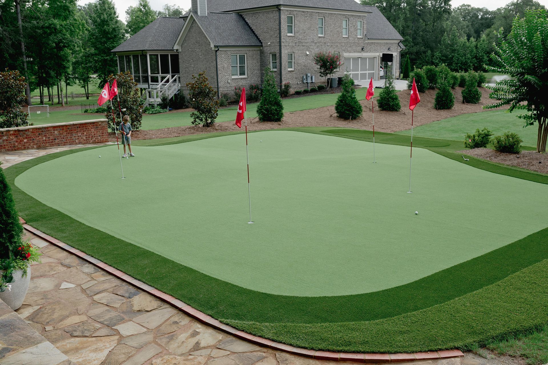 A backyard putting green with flags, surrounded by a dark green border and landscaping, in front of a brick home.