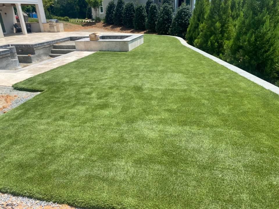 Lush green artificial turf in a backyard with a patio, raised planter, and evergreen trees.