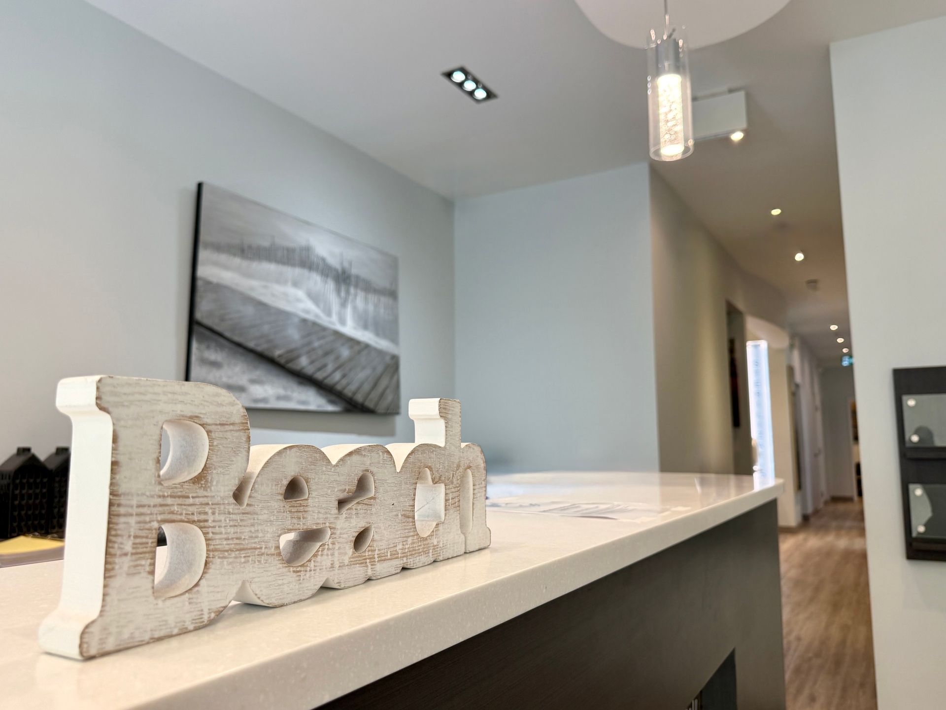 Modern kitchen with white island, gray walls, pendant light, and decorative “Beach” sign on the counter.