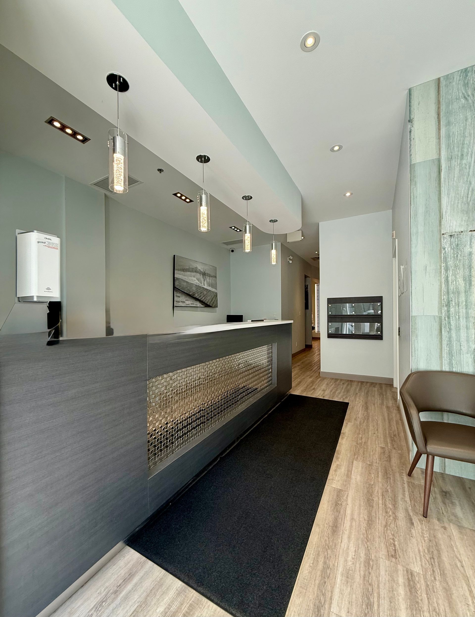 Modern office reception area with gray desk, black rug, wood floor, and hanging pendant lights.