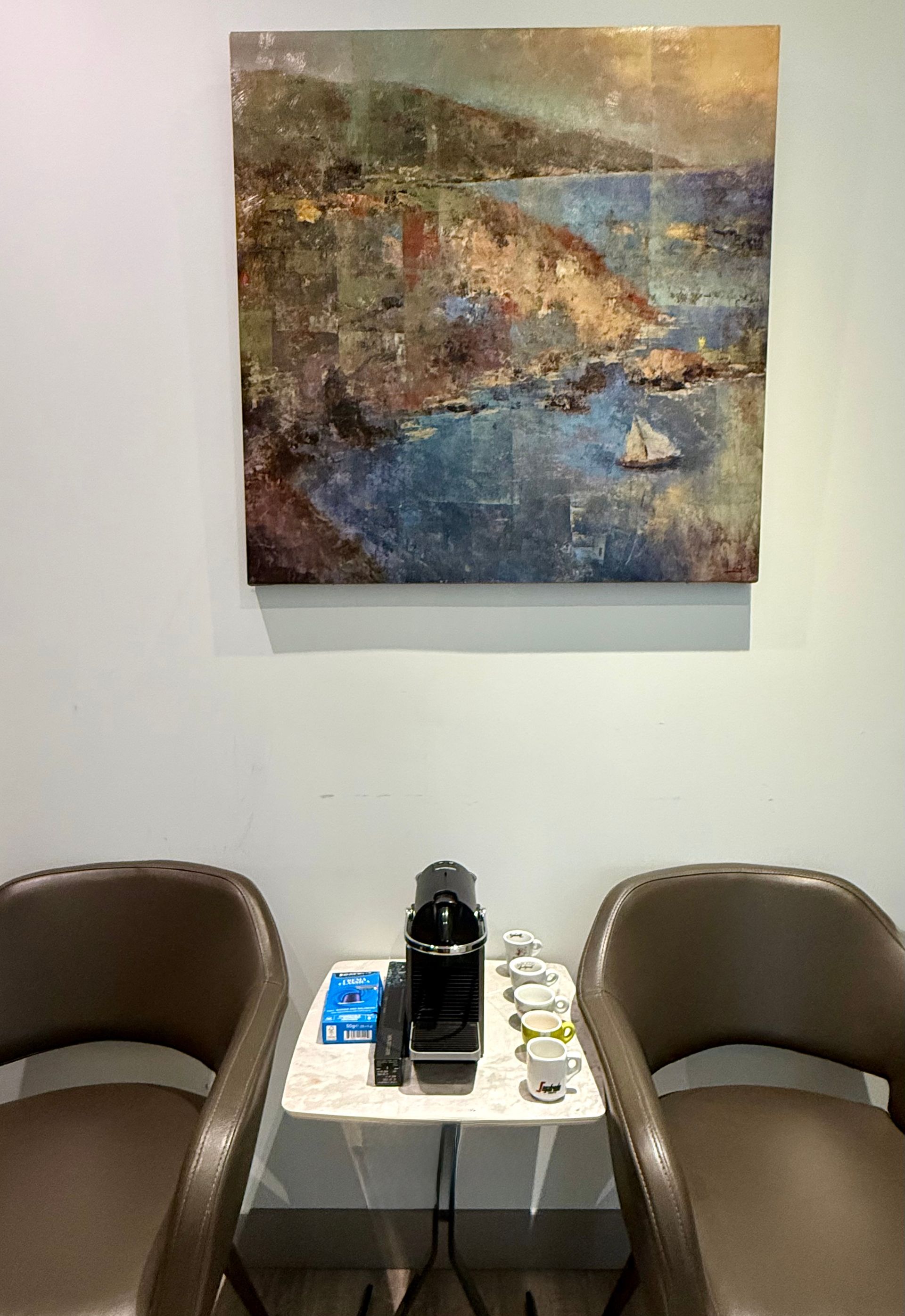 Two brown chairs flanking a small white table with a coffee maker under abstract wall art