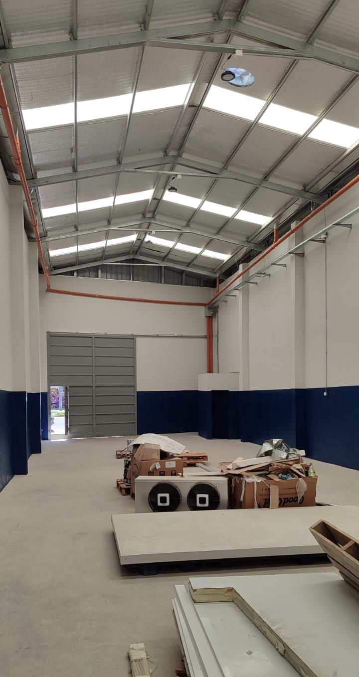 Interior vacío de un almacén con techos altos, tragaluces, paredes azules y blancas, y materiales de construcción en el suelo.