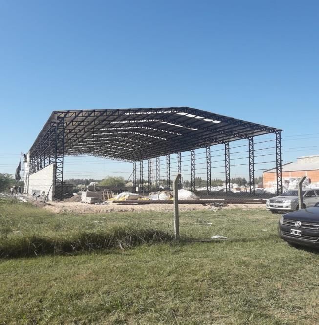 Edificio con estructura de acero en construcción sobre un campo de hierba, coches en primer plano, cielo azul despejado.