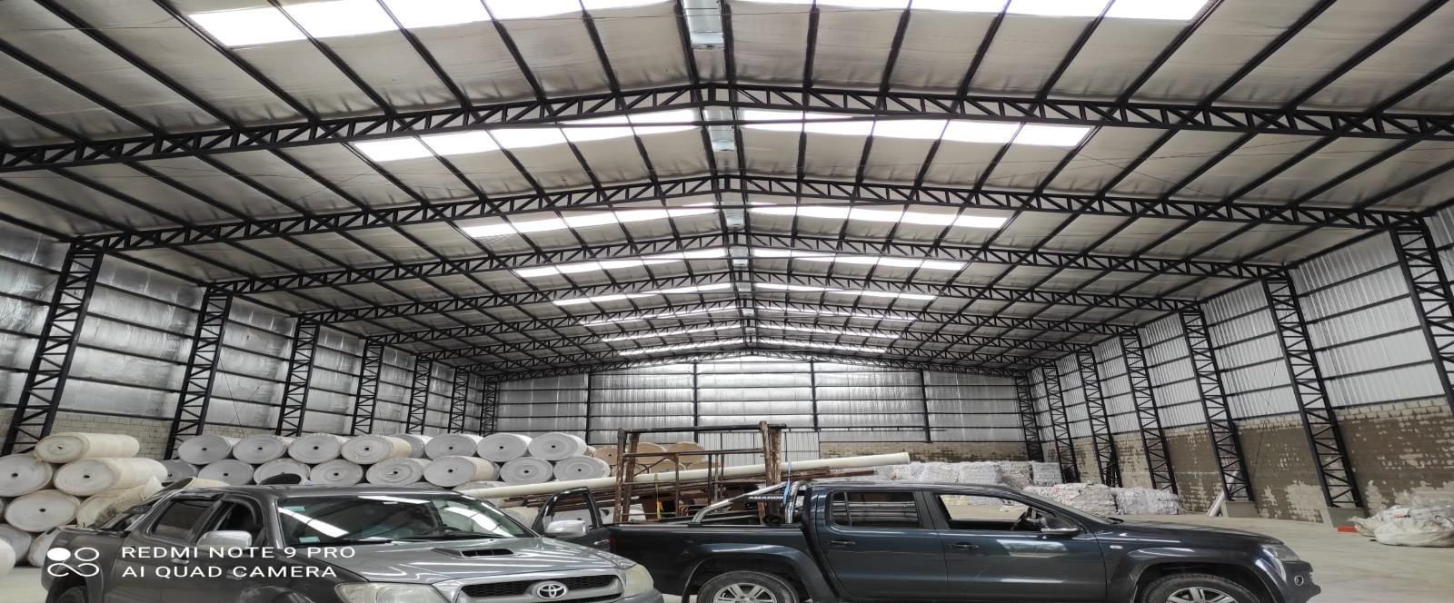 Dentro de un gran almacén con vehículos estacionados y materiales enrollados bajo una estructura de techo metálico.