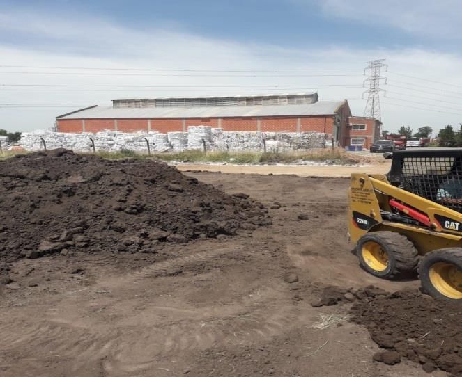 Una minicargadora en un terreno baldío con un montón de tierra oscura, un edificio industrial al fondo.