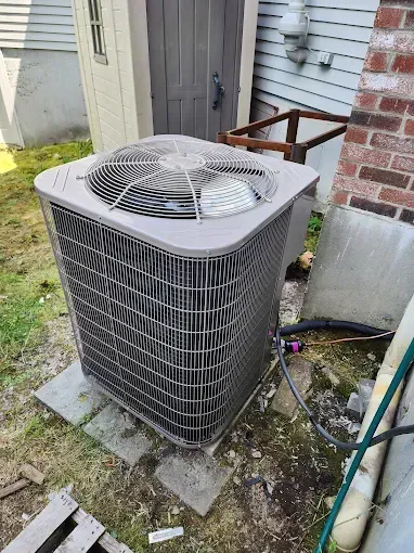 A gray outdoor HVAC condenser unit sits on concrete pavers next to a house wall and a storage shed.