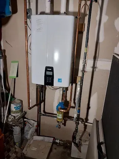 A wall-mounted white Navien tankless water heater with copper piping and a water filtration unit below it in a basement.