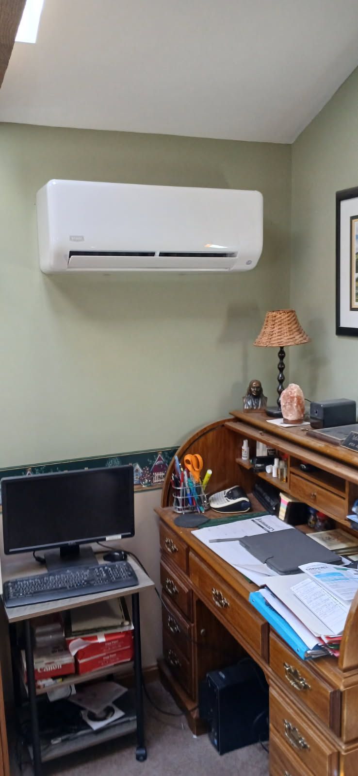 A wall-mounted air conditioner unit above a wooden roll-top desk and a computer workstation in a home office.