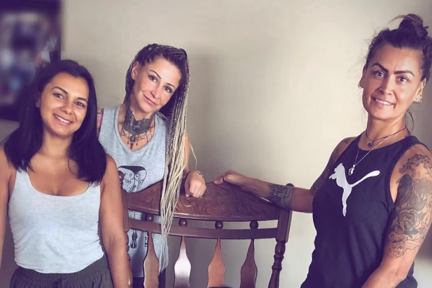 Three women smiling, posing near a wooden chair in a room. One has dreadlocks, another has arm tattoos.