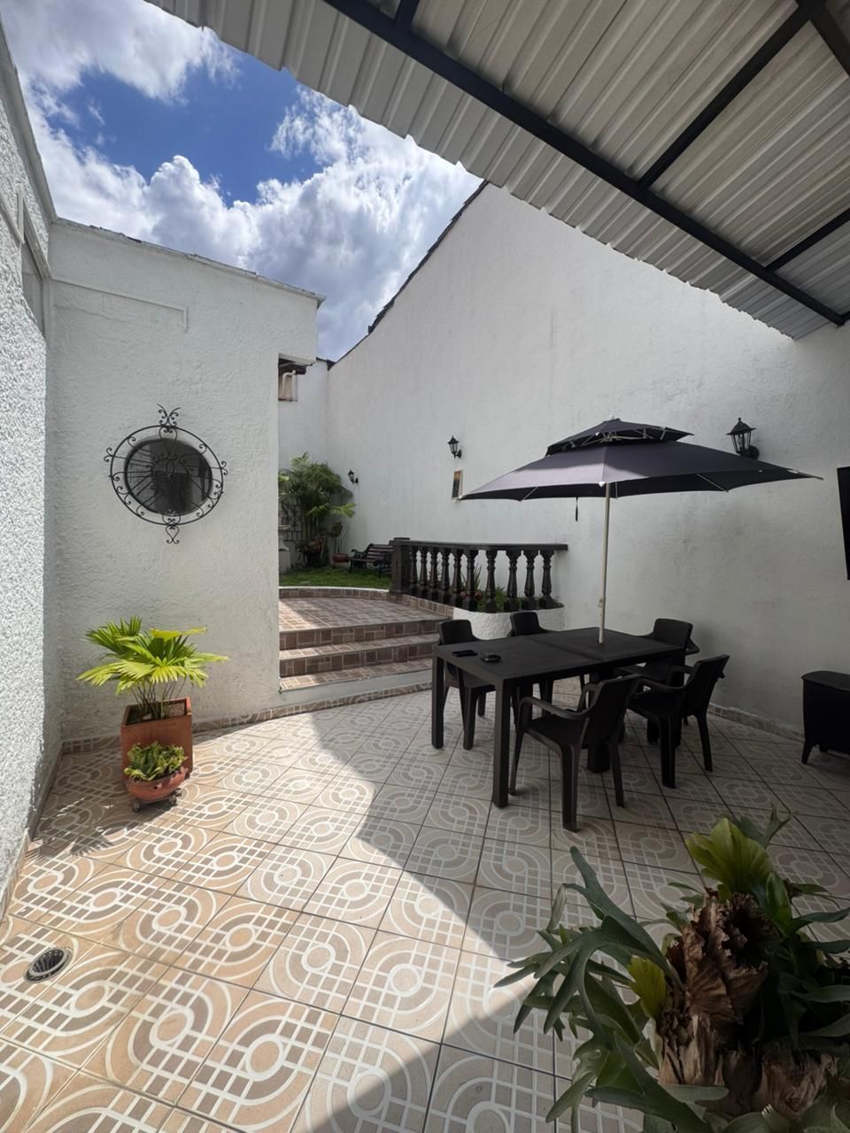 Patio soleado con suelo de baldosas, mesa y sombrilla negras, plantas en macetas y paredes de estuco blanco.