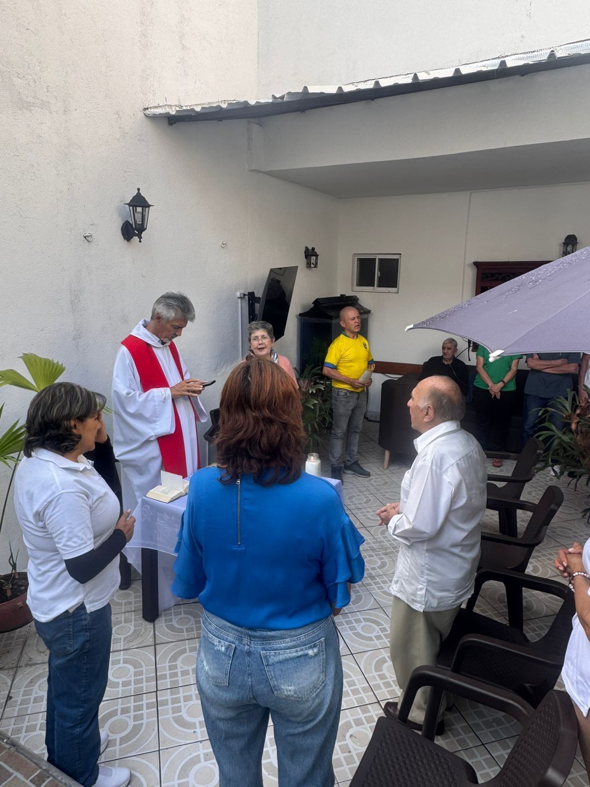 Grupo de personas charlando al aire libre cerca de un edificio blanco, mientras un sacerdote con ornamentos rojos y blancos pronuncia un discurso.