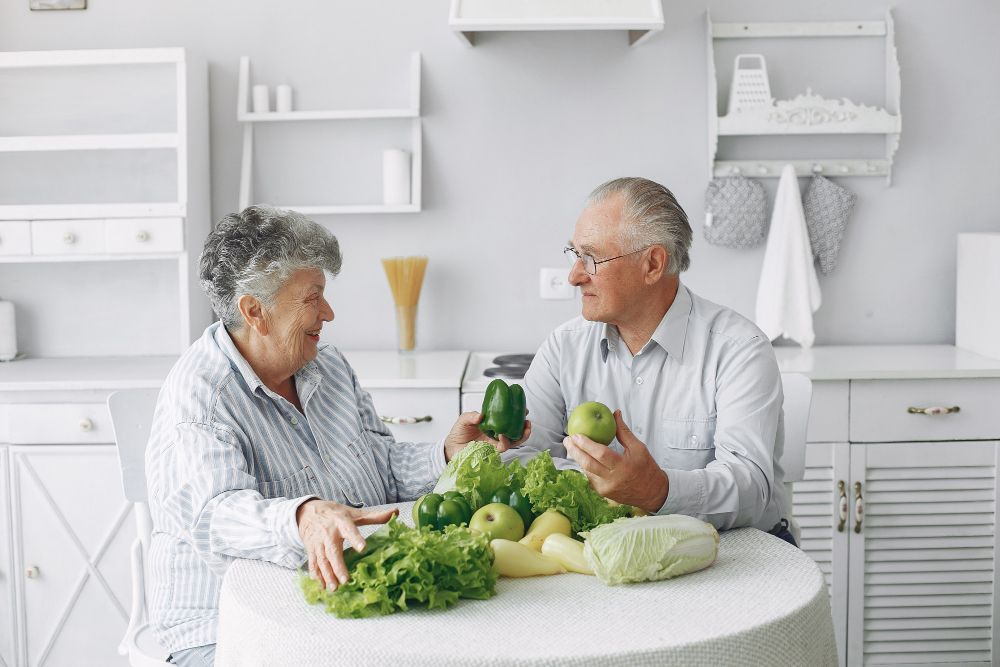 Dos personas charlando en una mesa de cocina con verduras frescas y una col, en una cocina de un blanco brillante.
