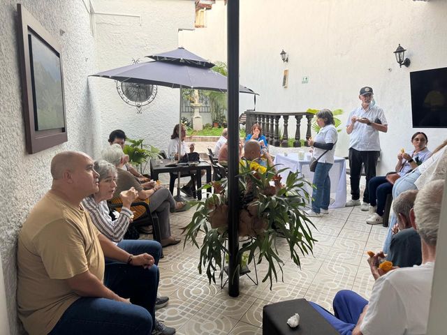 Varias personas sentadas en el patio de una cafetería, escuchando a un orador cerca de una pared con plantas y sombrillas.