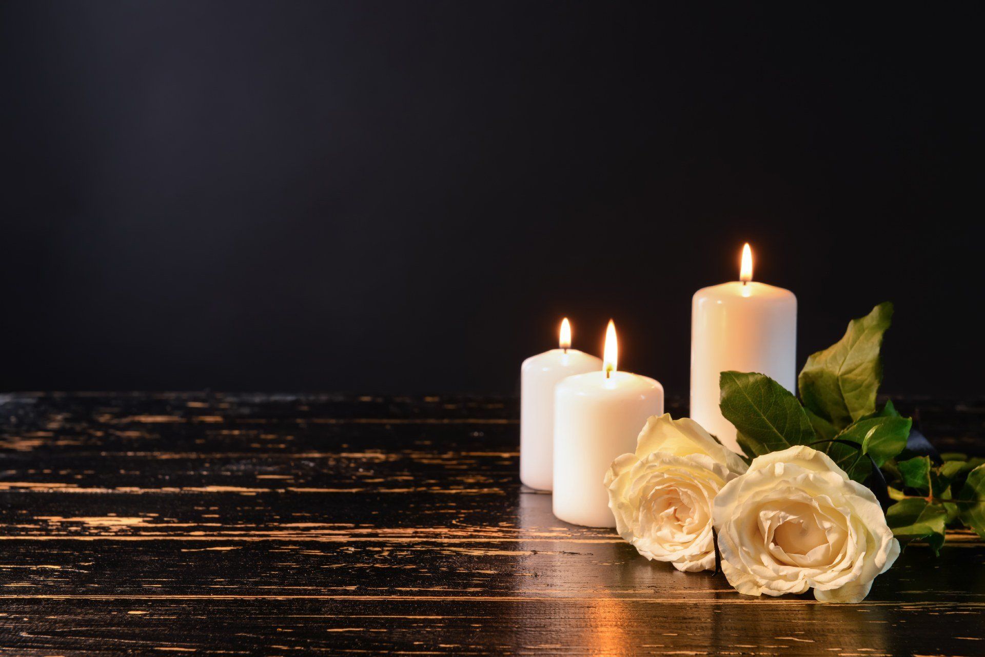Tre candele bianche accese e rose bianche su una superficie di legno nero. Sfondo scuro.
