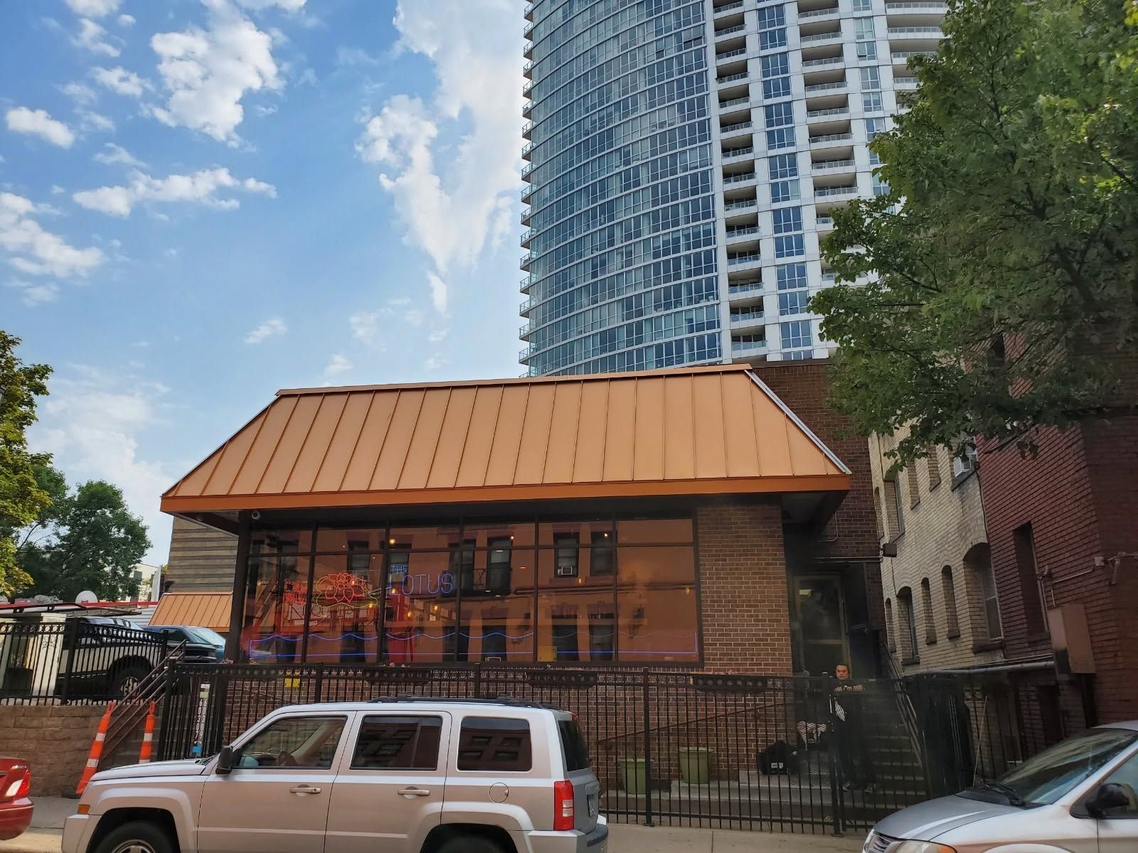 Brick building with copper roof behind a black fence, parked white SUV, and a tall apartment tower in back