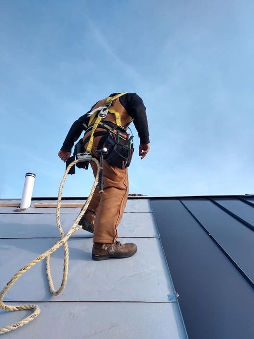 Worker on a roof wearing a safety harness, standing on metal roofing under a clear blue sky.