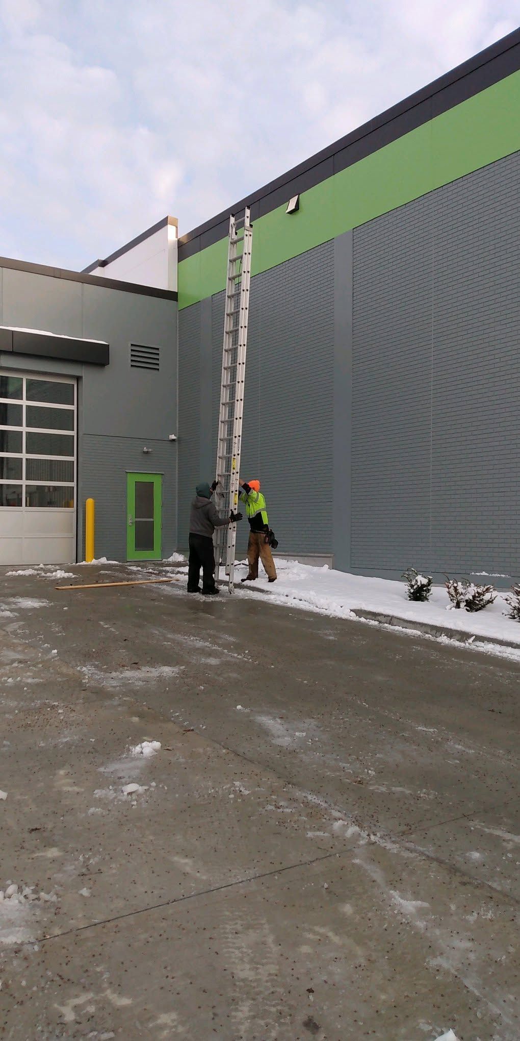 Two workers at a snowy industrial building, one on a ladder beside a tall gray wall with a green stripe.