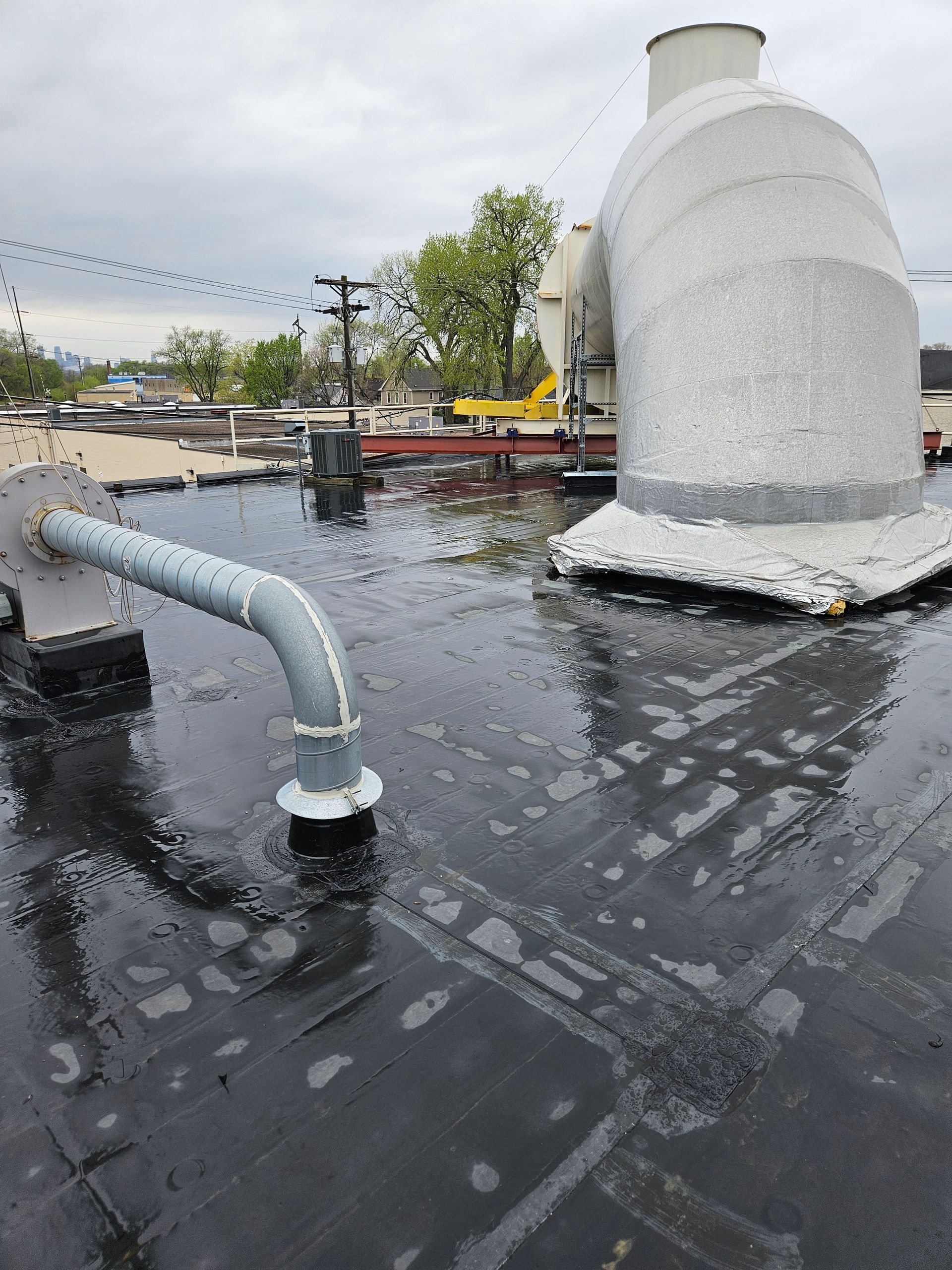 Rooftop with black membrane, gray vents, and puddled water after rain under an overcast sky
