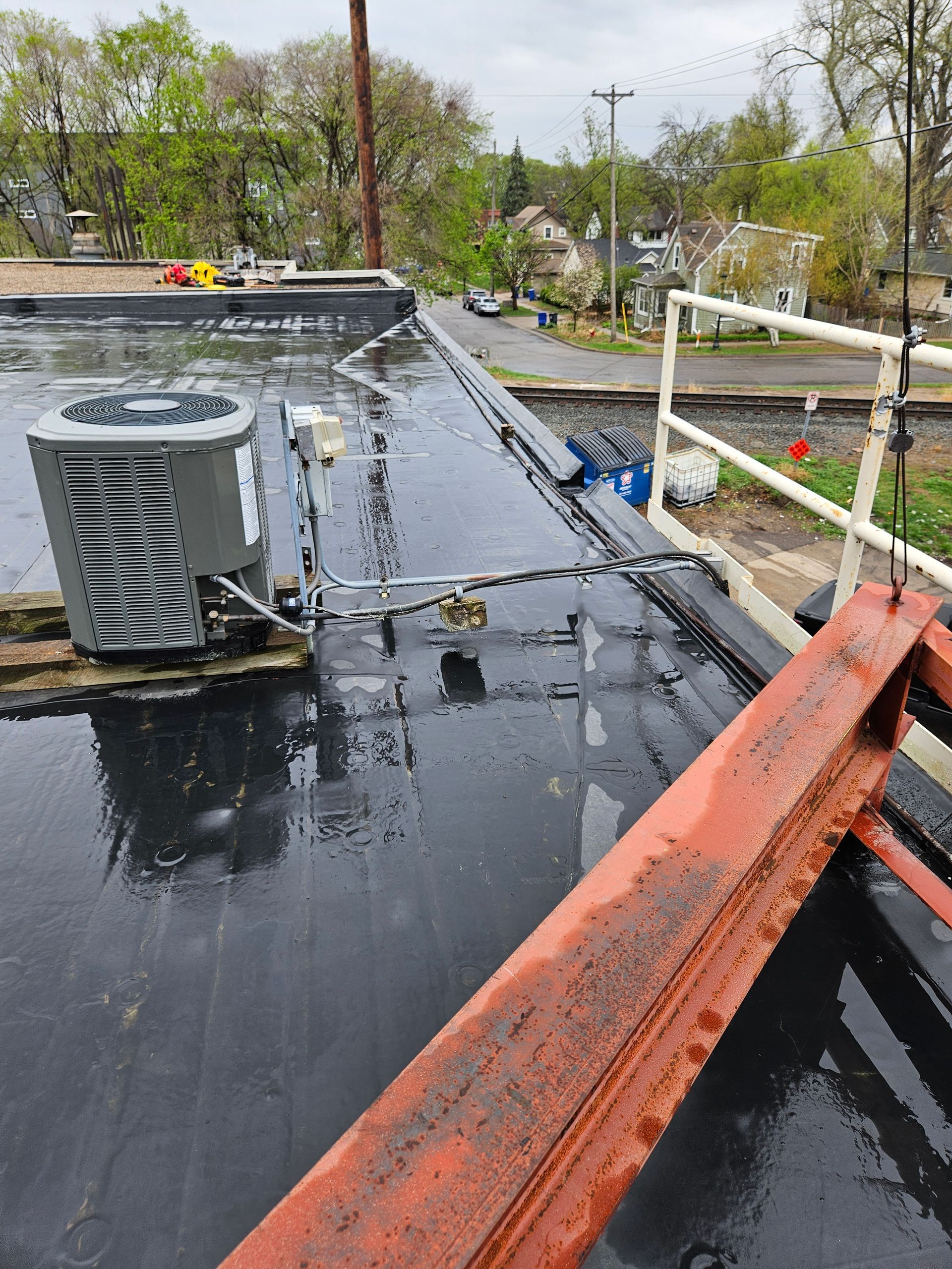 Rooftop with wet black surface, HVAC unit, and orange safety railings under a cloudy sky.