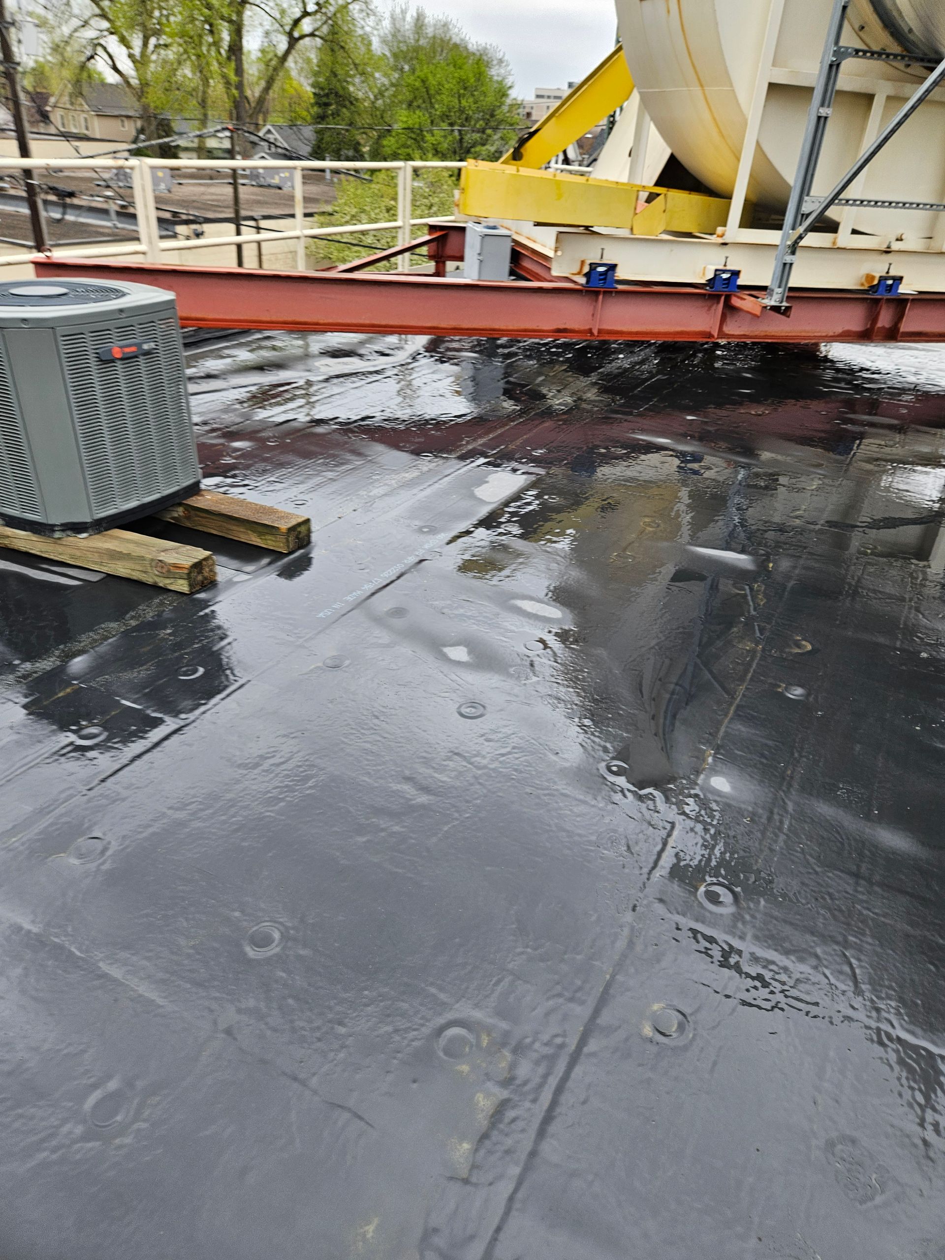 Rooftop with dark waterproof membrane, metal equipment, and a yellow concrete mixer under cloudy skies