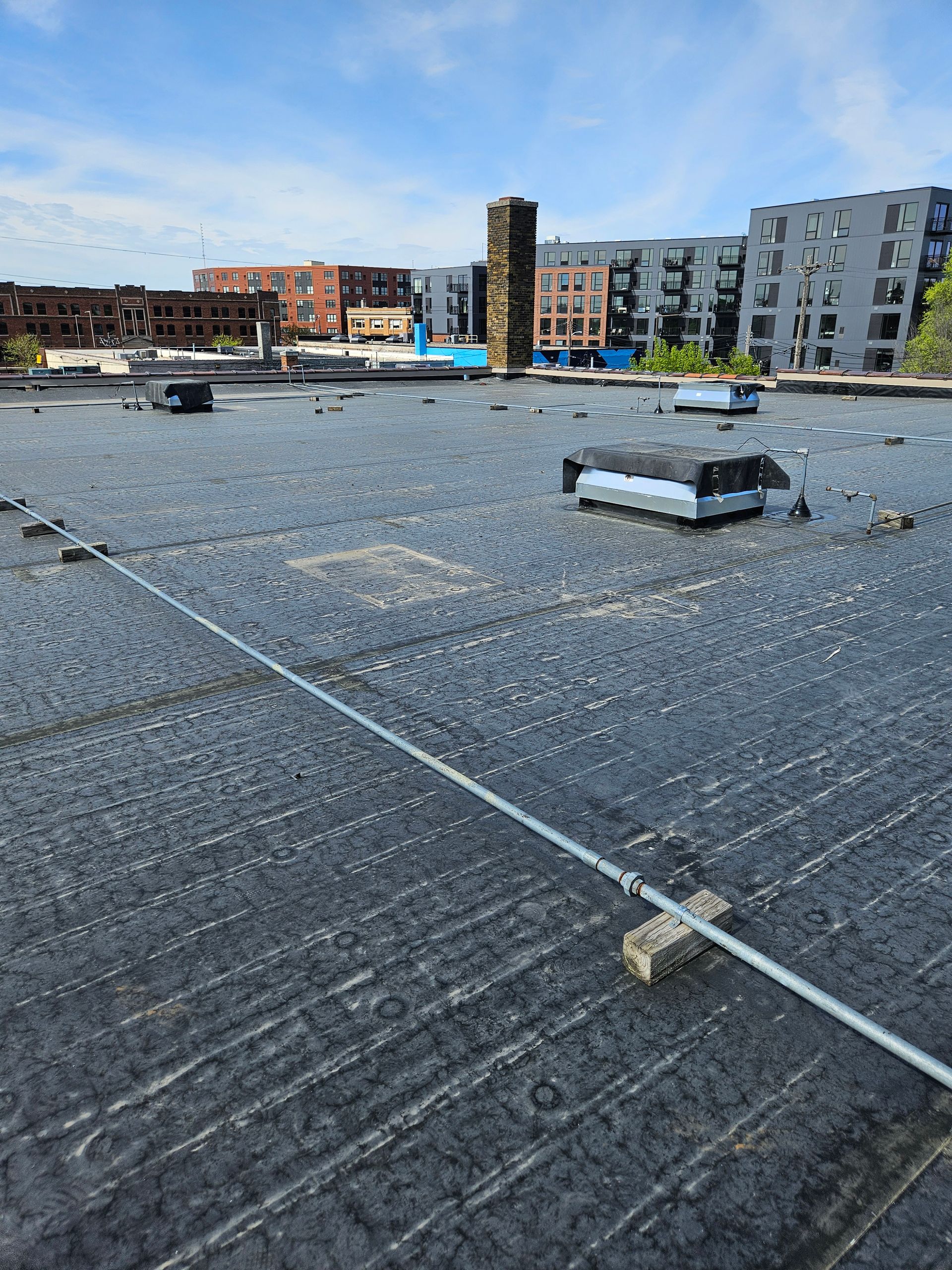 Rooftop with black membrane, vents, and city buildings under a blue sky