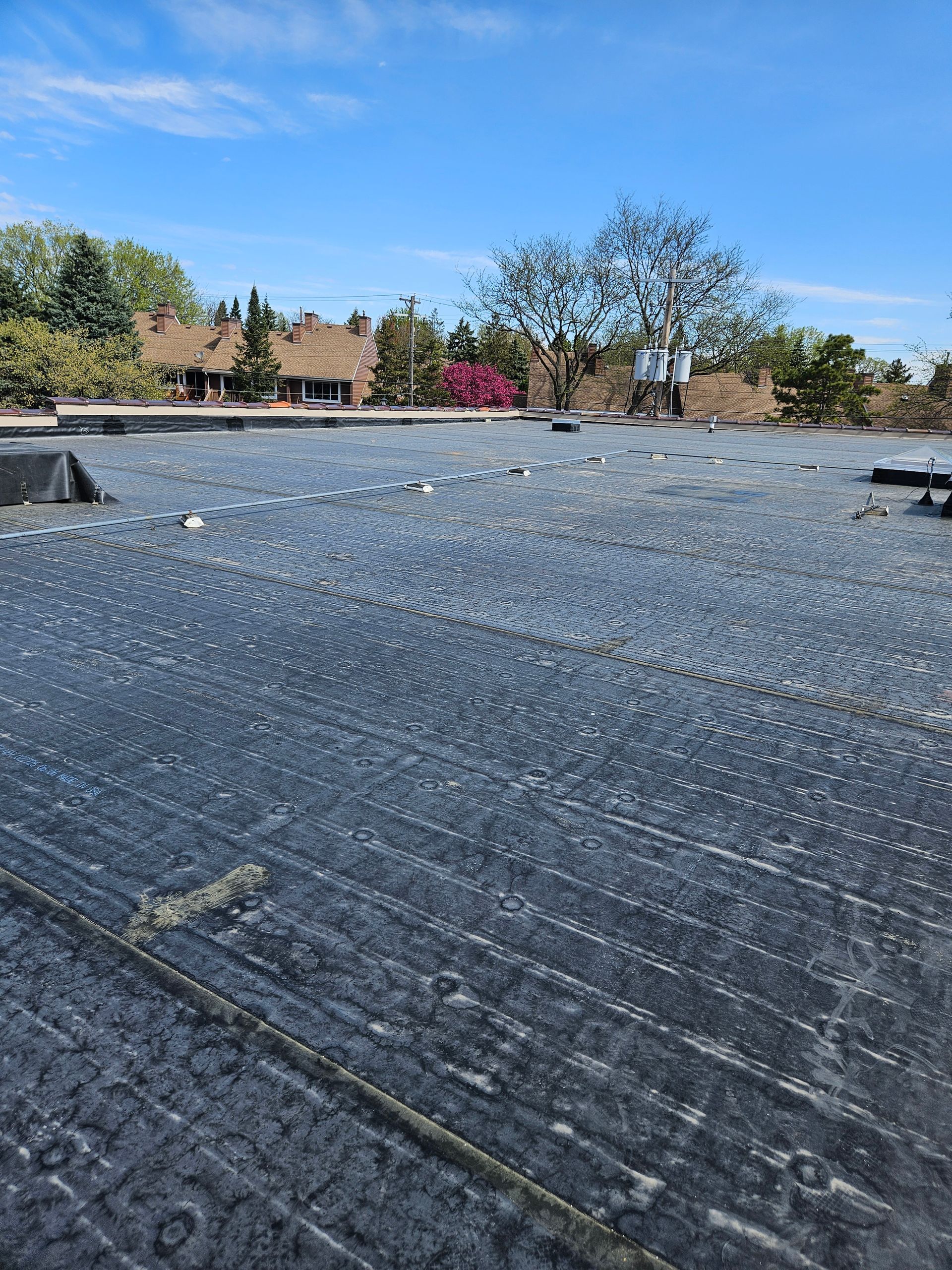 Black tarped rooftop with white debris under a clear blue sky, trees and houses in the background