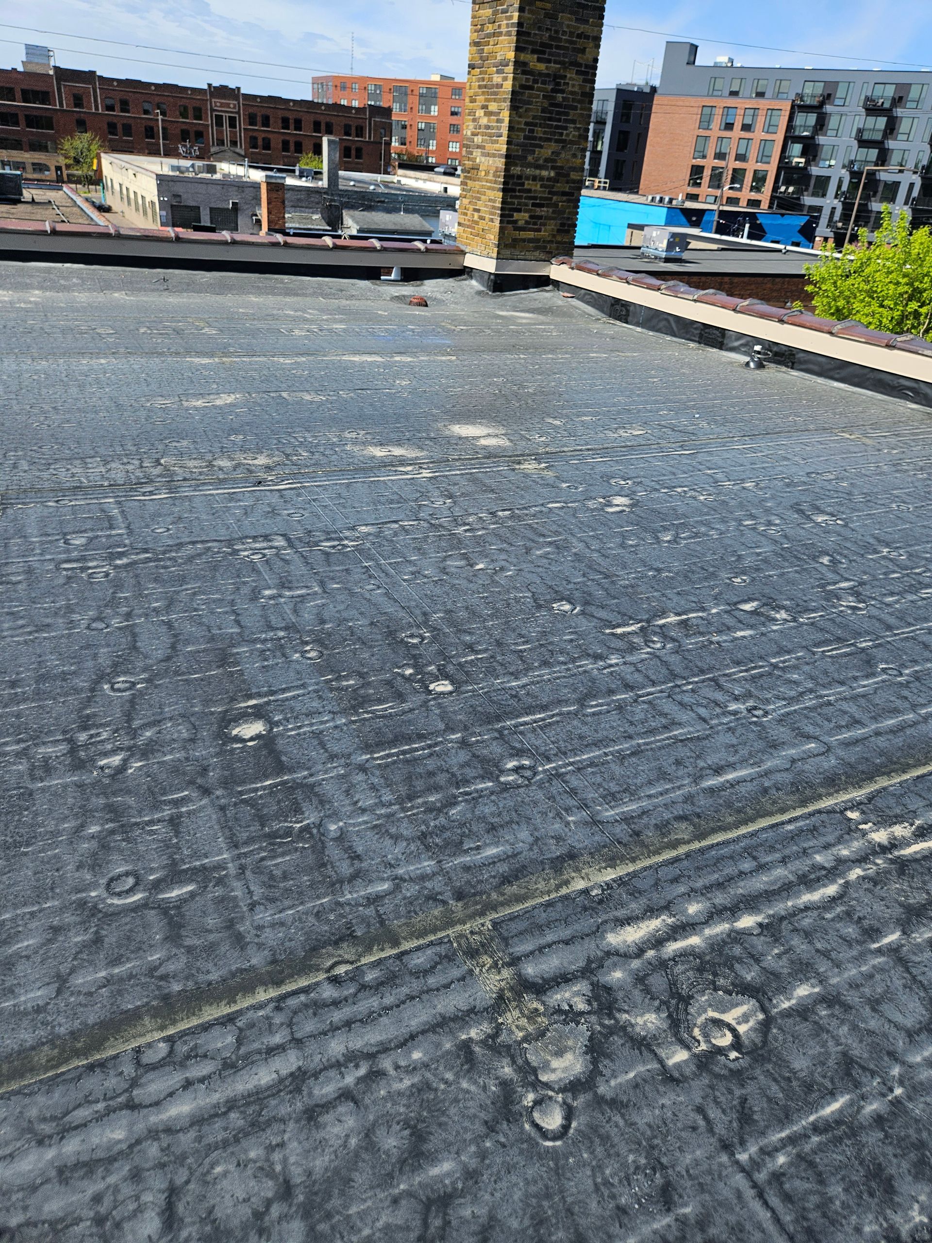 Rooftop covered in black roofing membrane with a brick chimney and city buildings in the background