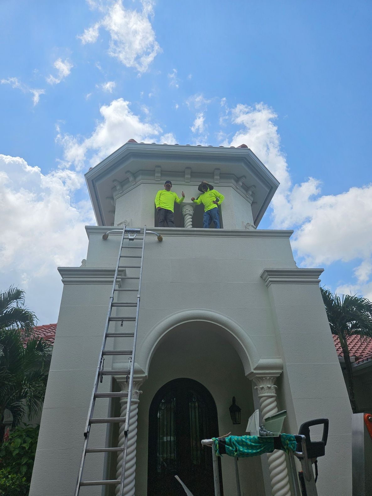 Two workers in neon shirts on a white tower, one on a ladder under a blue sky with clouds