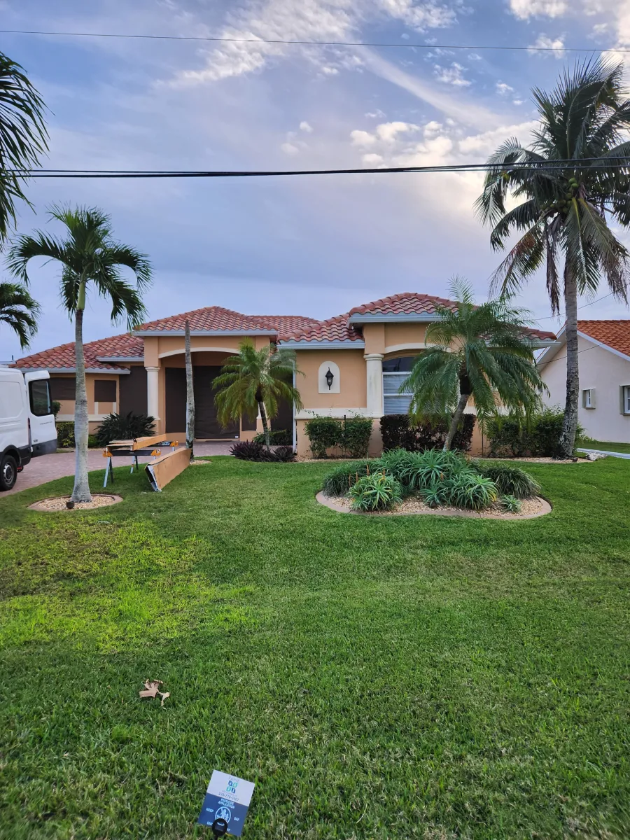 Single-story house with red-tile roof, palm trees, and a green lawn under a cloudy sky