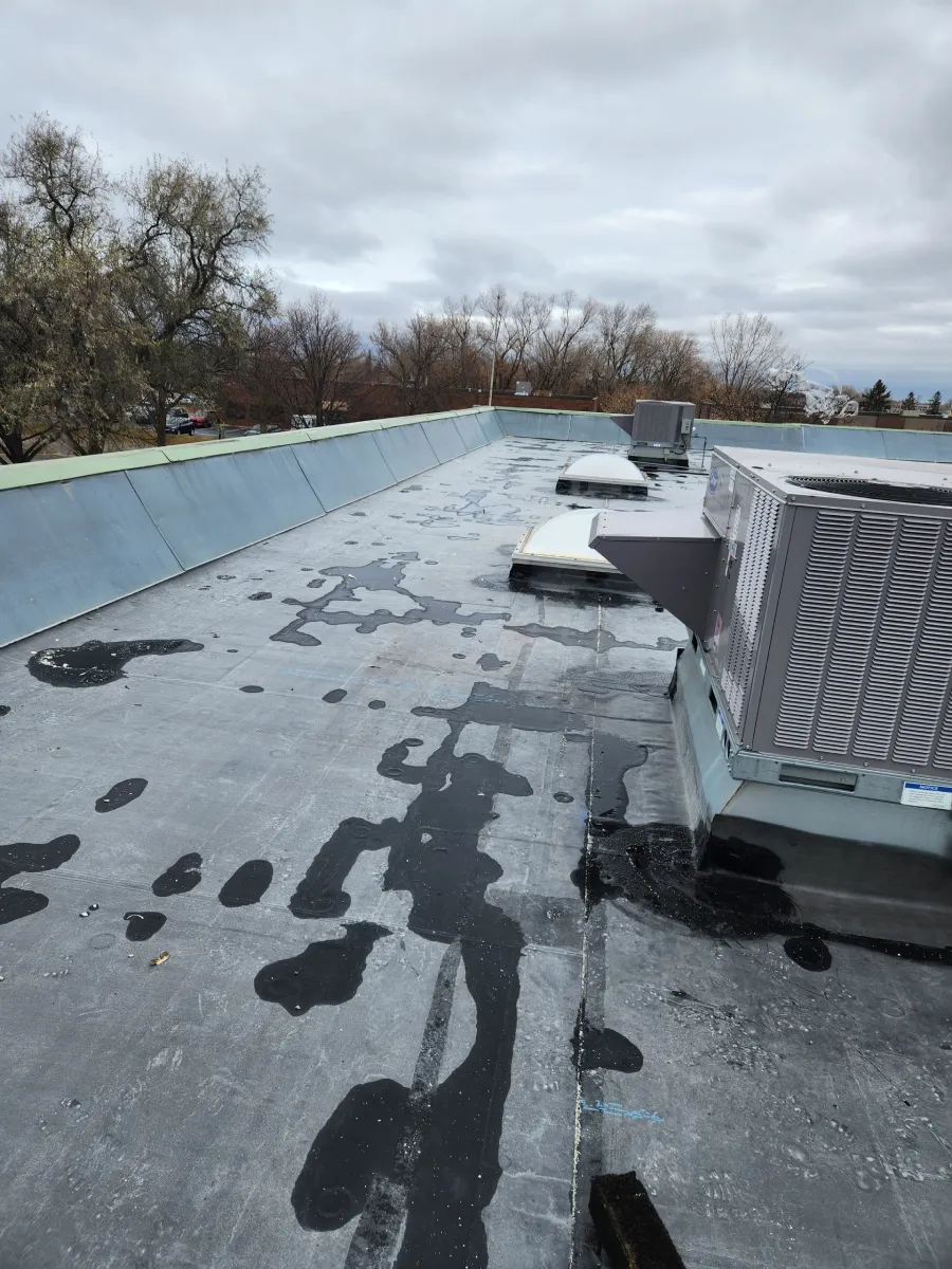 Flat commercial rooftop with HVAC units, vents, and patchy dark waterproofing under a cloudy sky